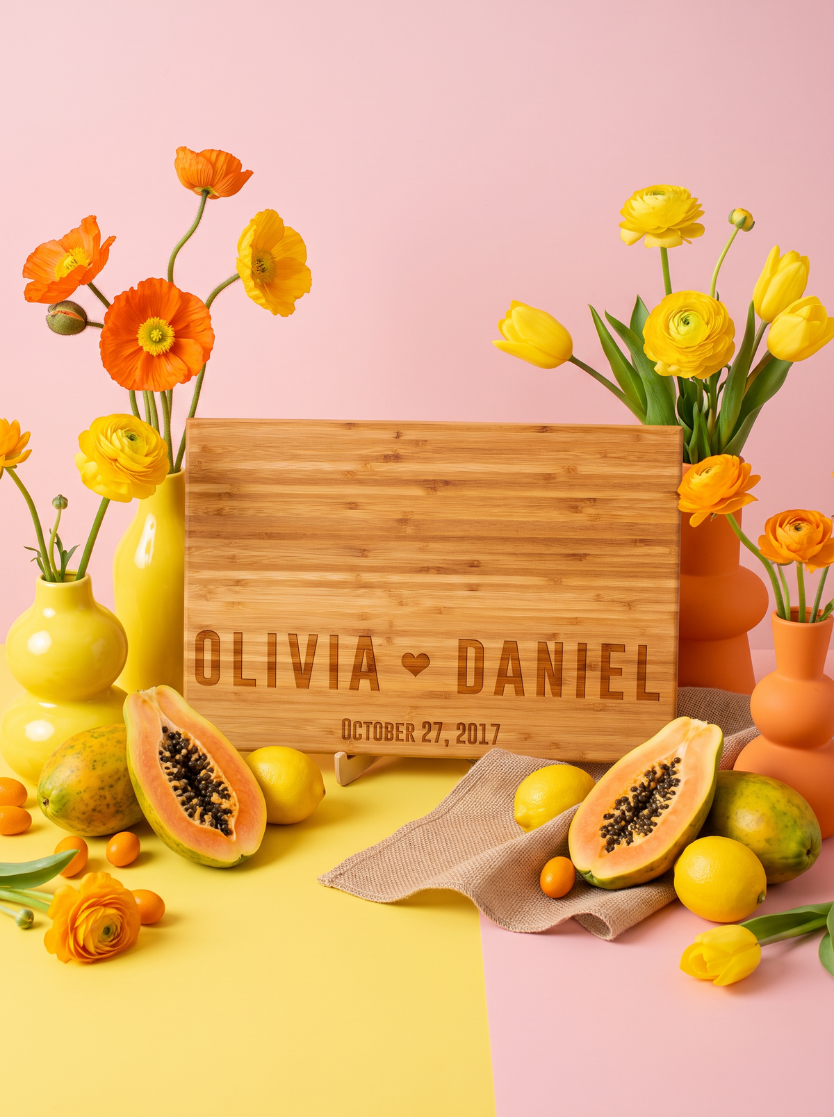 Bamboo cutting board engraved with two bold names, a heart and date, styled with yellow flowers on pink.