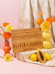 Angled view of a bamboo cutting board engraved with two bold names, a heart and date, beside yellow and orange flowers.