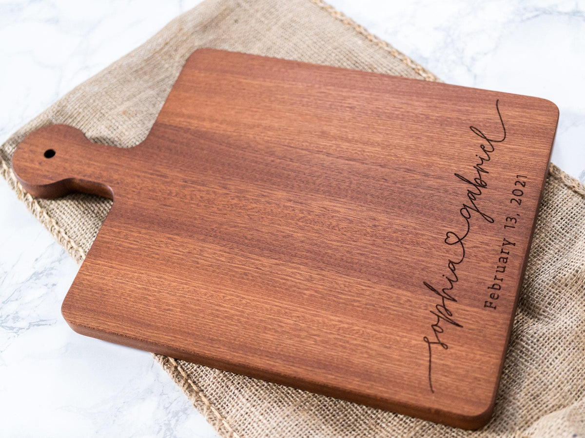 A rectangular wooden serving board with rounded corners and a handle, shown on a burlap cloth over a light marble surface. The board is engraved with the names 'Sophia & Gabriel' and the date 'February 13, 2021' in dark script.