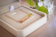A gold-colored necklace with a rectangular pendant is displayed in a beige jewelry box, accompanied by a bouquet of white flowers.