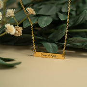 A gold-colored necklace with a rectangular pendant engraved with the names "Eve & Ada". The necklace is displayed against a backdrop of green leaves and white flowers.