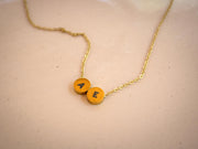 A gold chain necklace with two round yellow-orange initial pendants spelling 'AE', resting on a light beige textured surface.