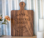 Holly Mixing Bowl edited mockup on a large dark wooden paddle board, showing Nana’s Christmas Bakery and grandkids’ names against striped wallpaper.