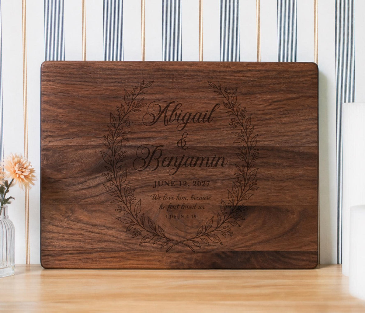 Edited mockup of first loved wreath on a horizontal rectangular board with a leafy wreath, two names, date and 1 John 4:19.