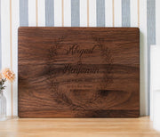 Edited mockup of first loved wreath on a horizontal rectangular board with a leafy wreath, two names, date and 1 John 4:19.