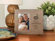 Taupe-colored frame beside a lamp and plant, showing a nest design, “Our first GRANDCHILD,” sample Oliver / JAMES / 2026, and an illustrative portrait of grandparents holding a newborn.