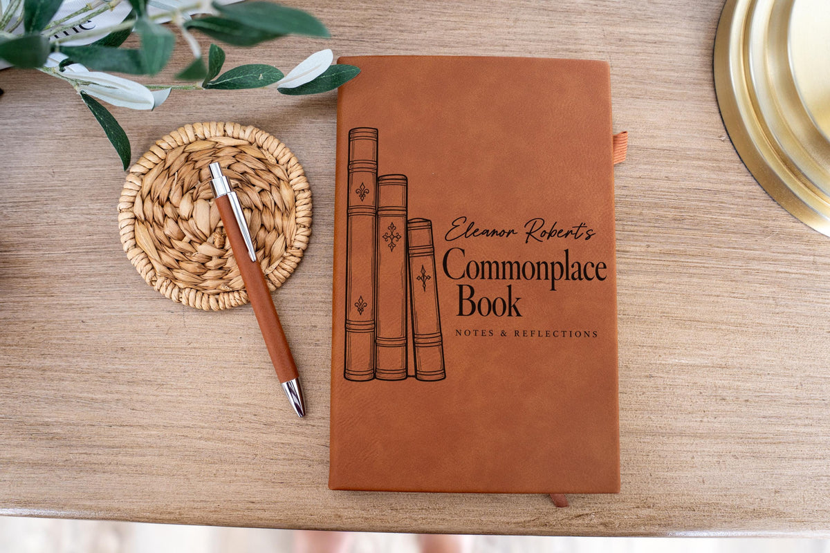 Edited mockup of a brown Commonplace Book journal on a light wood surface, with a pen on a woven coaster beside it; sample name Eleanor Robert’s above the title.