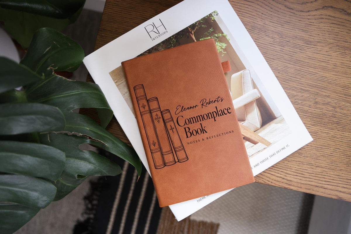 Angled edited mockup of a brown Commonplace Book journal resting on a white book beside green leaves; three illustrated book spines, sample name Eleanor Robert’s and Notes & Reflections.