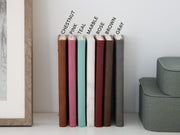 Seven journals standing on a shelf, labeled Chestnut, Pink, Teal, Marble, Rose, Brown and Gray, beside stacked green cases.