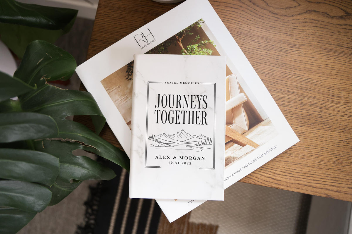 Edited mockup of a marble-pattern journal with Journeys Together lettering, mountains, river and pine trees, personalized Alex & Morgan and 12.31.2025, on a magazine beside green leaves.