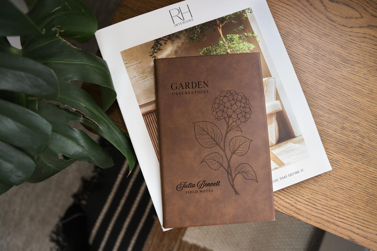 Brown Garden Hydrangea journal mockup on a book beside green leaves and a wood table. Cover shows a hydrangea, Garden Observations, Julia Bennett, and Field Notes.