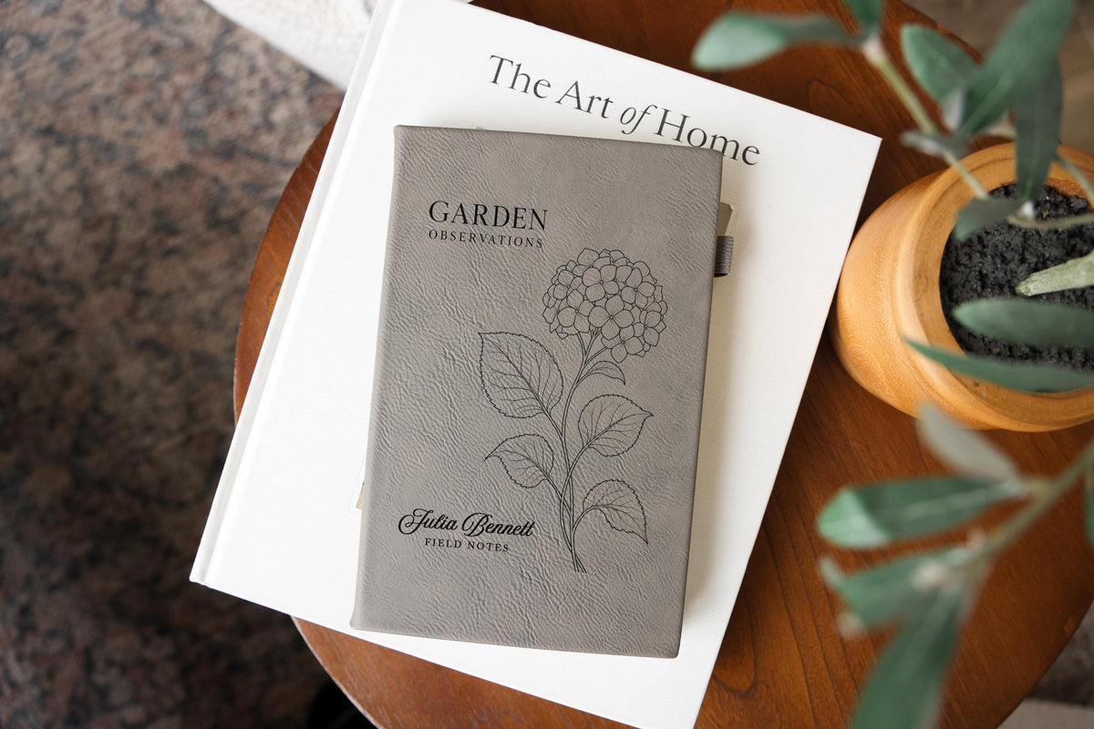 Gray Garden Hydrangea journal mockup on a book beside a potted plant. Cover shows a hydrangea, Garden Observations, Julia Bennett, and Field Notes.
