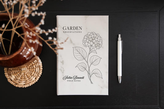 Marble Garden Hydrangea journal mockup on a dark table beside a white pen and dried flowers. Cover shows a hydrangea, Garden Observations, Julia Bennett, and Field Notes.
