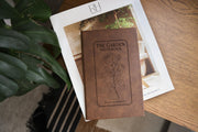 Brown The Garden journal mockup on a book beside broad green leaves and a wood table. Cover shows a rose stem, a fine border, The Garden Notebook, and the sample name Julia Bennett.