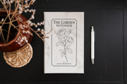 Marble The Garden journal mockup on a dark table beside a white pen and dried flowers. Cover shows a rose stem, a fine border, The Garden Notebook, and the sample name Julia Bennett.