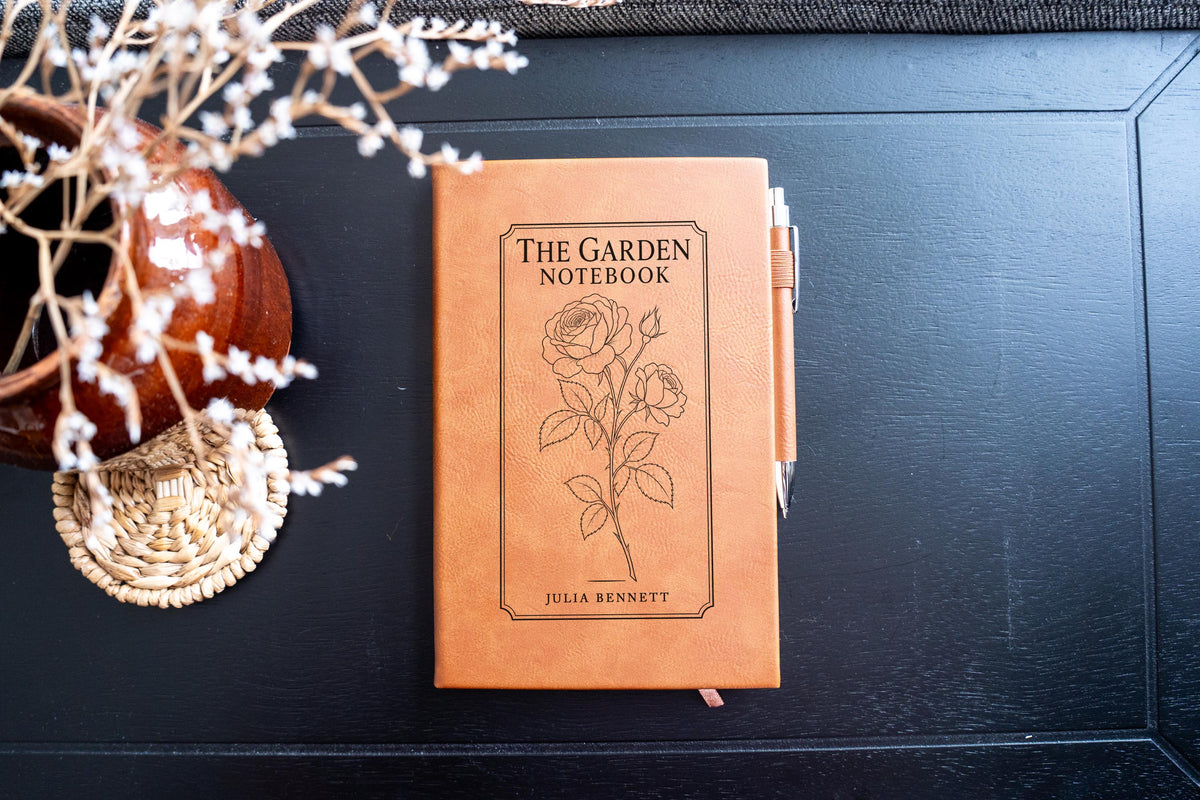 Chestnut The Garden journal mockup on a dark table with a matching pen and dried flowers. Cover shows a rose stem, a fine border, The Garden Notebook, and the sample name Julia Bennett.