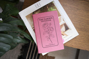Pink The Garden journal mockup on a book beside broad green leaves and a wood table. Cover shows a rose stem, a fine border, The Garden Notebook, and the sample name Julia Bennett.