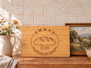 Edited mockup of a bamboo rectangle cutting board with Emma’s Sourdough Bakery loaf emblem and EST. 2020, styled on a kitchen counter.
