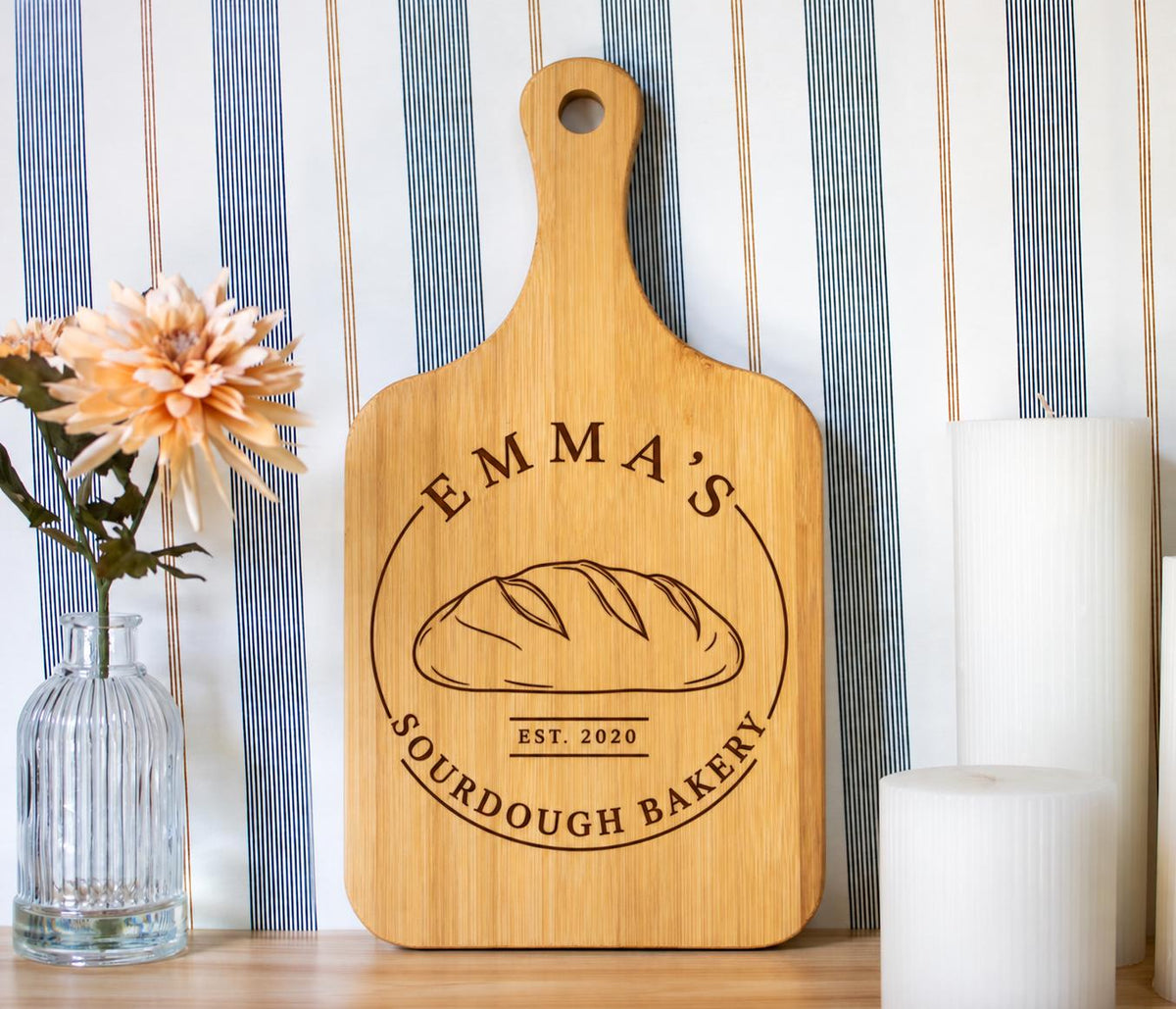 Edited mockup of a bamboo board with a handle, showing Emma’s Sourdough Bakery engraving beside flowers and candles.