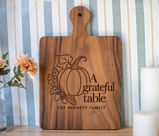 Grateful Table pumpkin, oak leaves and Bennett family engraving mockup on a walnut paddle board beside flowers and candles.
