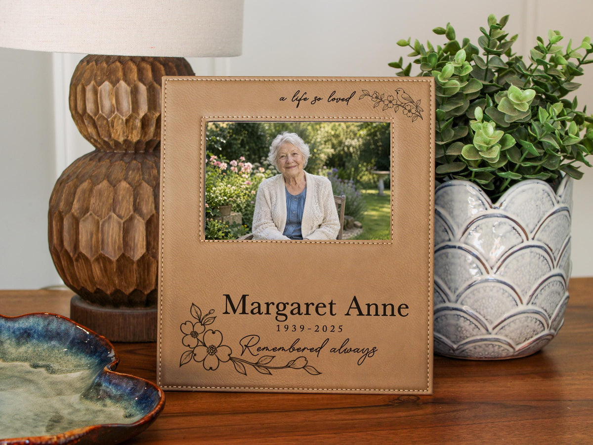Edited memorial frame mockup beside a lamp and plant, with a life so loved songbird engraving and an illustrative elderly woman's portrait above Margaret Anne and dates.