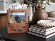 Edited mockup of the Pacific Crest Trail frame on a dark table beside books and a vase, with mountain artwork and an illustrative hiking couple photo.