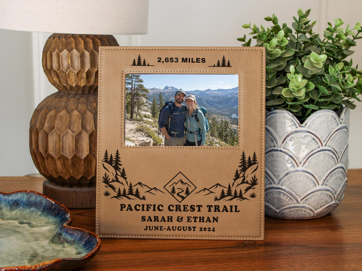 Edited mockup of the Pacific Crest Trail frame beside a lamp and plant, with mileage, mountain and pine artwork, names, date and an illustrative hiking couple photo.
