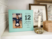 Edited teal Varsity Volleyball frame mockup on a shelf beside a candle and framed art, with an illustrative player portrait and personalized varsity number, name, school and year.