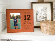 Warm reddish-brown Varsity Field Hockey frame on a white shelf, with an illustrative player portrait, number 12, Jordan Miller, crossed field hockey sticks and 2026.