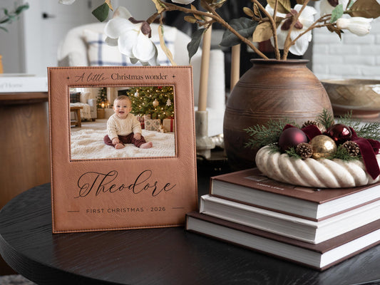 Edited mockup of Christmas Wonder frame engraved with Theodore and First Christmas 2026, an illustrative baby photo, and festive greenery and ornaments beside it.