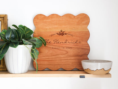 a wooden cutting board with the name "The Hendricks" and the date "August 13, 2025" engraved on it, placed on a shelf next to a potted plant and a ceramic bowl.