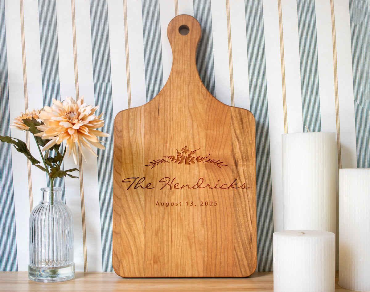 a wooden cutting board with the name "The Hendricks" engraved on it, placed on a table next to a vase with flowers and candles.