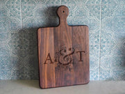 A wooden cutting board with the initials 'A&T' engraved on it, placed against a patterned tile wall.