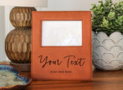 a brown leather picture frame with a blank white space in the center, placed on a wooden surface. The frame has a personalized message that reads "Your Text" and "your text here". In the background, there are decorative items such as a lamp and a potted plant.