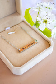 A silver bar necklace engraved with 'love over fear' in black script, presented in a beige velvet jewelry box with white flowers in the background.
