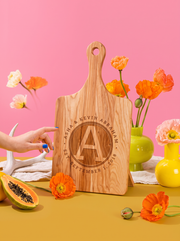 Wood paddle cutting board with a circular initial monogram, names and date engraving, styled with colorful flowers, view 2.