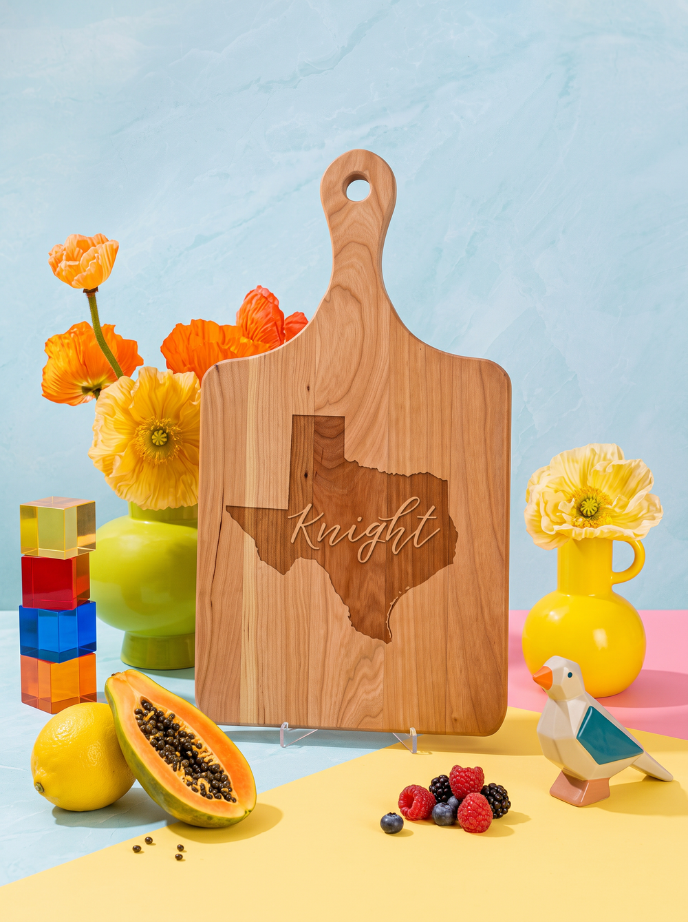 Wood paddle cutting board engraved with the Texas outline and a family name, styled with bright flowers against a blue background.